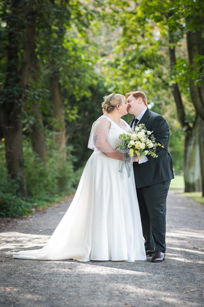 Das Brautpaar steht in einer Allee und küsst sich